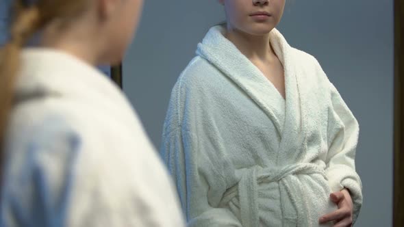 Pregnant Young Woman in Bathrobe Looking at Tummy in Mirror, Baby Expectation