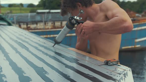 Topless young man using manual sealant gun to seal cabin roof planks on wooden boat, MID SHOT