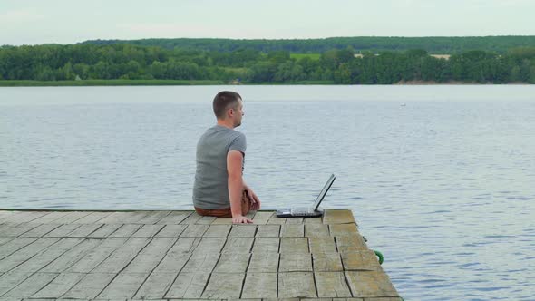 Handsome man sits on a wooden pier with laptop. Work on vacation