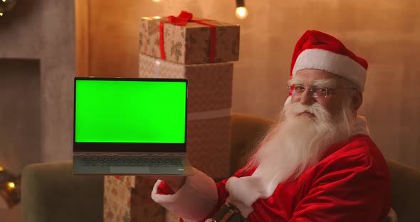 In the Background of the Christmas Tree, an Elderly Santa Claus Is Holding a Laptop and Pointing at