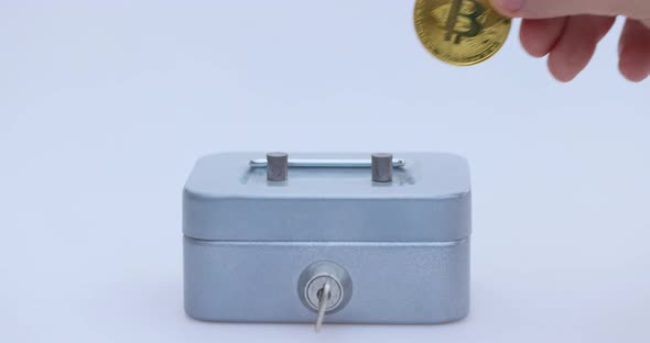 Male Hand Puts a Cryptocoin Bitcoin or BTC in a Small Grey Vault Safe or Piggy Bank Depicting Crypto