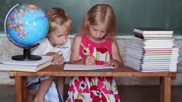 A Brother Sits Down To His Sister Who Is Drawing for School. Home School Concept.