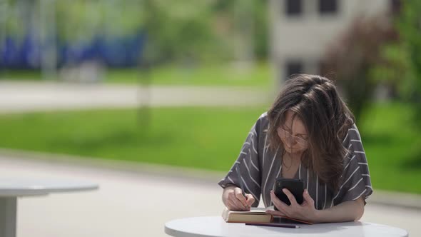 Smart Woman is Reading Book and Using Smartphone for Translating Making Notes on Page