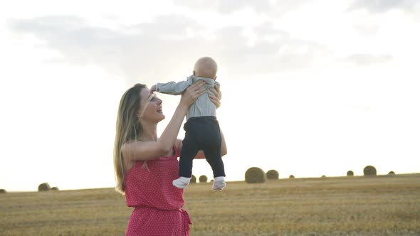 Happy Mother Throwing Her Baby Son in a Field Near Haystacks on Sky Background