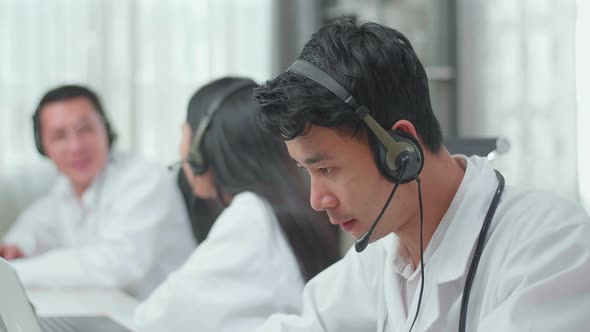 Asian Man Doctor Working As Call Centre Agent Speaking To Customer While His Colleagues Are Talking