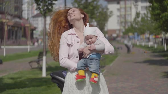 Young Beautiful Redhead Mother Holding a Cute Baby Boy in Her Arms and Talking To Him in a Spring