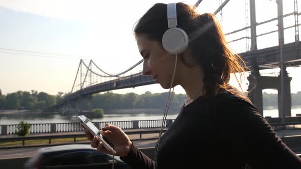 Beautiful Girl in White Headphones Listens To Music and Looks Into the Phone. Pedestrian Bridge