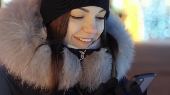 Young Beautiful Woman Uses the Phone Against the Background of the Central Christmas Tree Lights and