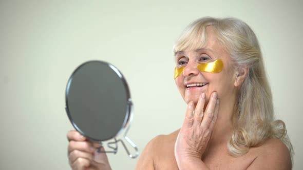 Joyful Smiling Lady Applying Eye Patches Looking in Mirror, Anti-Aging Therapy