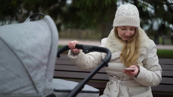 Happy Mother with Smartphone Using App. She Sitting on the Bench , Baby Sleeping in Pram