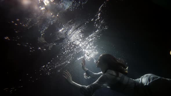 A Beautiful Woman in a Flowing Dress Floats Under the Water on a Dark Background She Swims To the