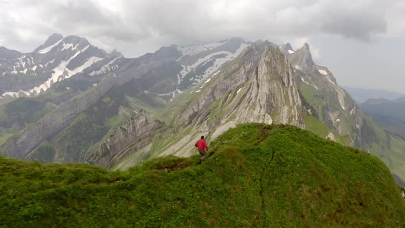 A man running on top of a mountain. He is wearing a red jacket. To his left and right it goes down a