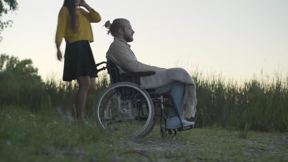 Side View Wide Shot of Paralyzed Man on Wheelchair Resting with Girlfriend Outdoors at Dusk