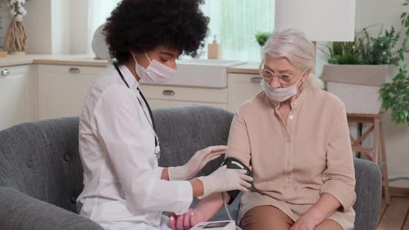 Afro American Woman Doctor in Mask Checks Blood Pressure Senier Woman at Home Sitting on Couch