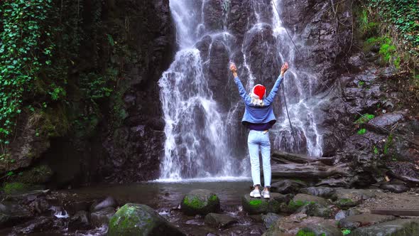 A girl in a Santa Claus hat stands on a waterfall with her arms outstretched.