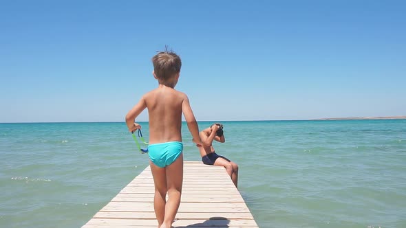 Little Boy in Swimming Trunks Goes To His Brother, a Teenager on a Wooden Pier on a Sunny Hot Day To