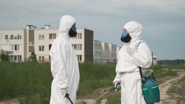 Two people in respirators and protective suits are talking while standing in field