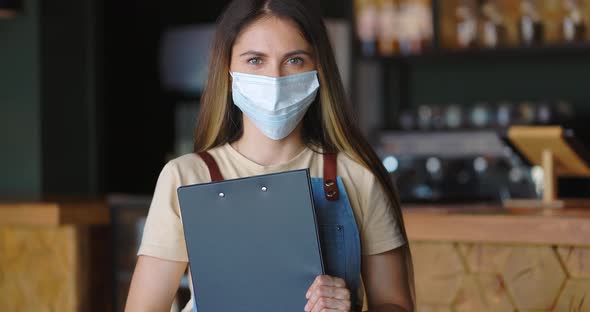Friendly Waitress in Protective Mask Posing at Cafe with Menu in Hands