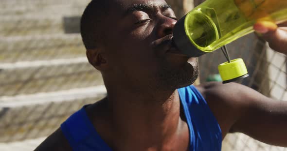 Tired african american man sitting and drinking from water bottle, taking break in exercise outdoors