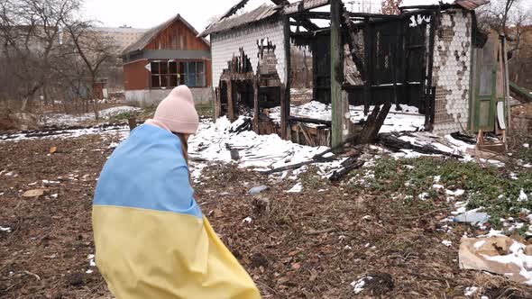 A sad girl wearing in the Ukrainian flag walks against the background of destroyed houses. War in Uk