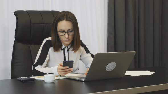 Woman Works on Laptop in Office with Phone in Her Hand