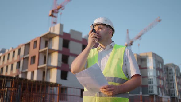 Foreman in Helmet and Vest Talking on Walkietalkie with Builders Standing at Construction Site