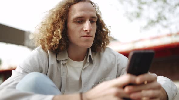 Pensive curly-haired man typing by phone