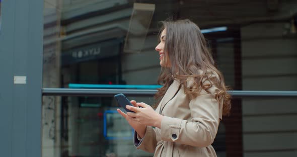 Attractive Young Woman Using Phone Walking Near Big Modern Office Building. Chatting with Friends