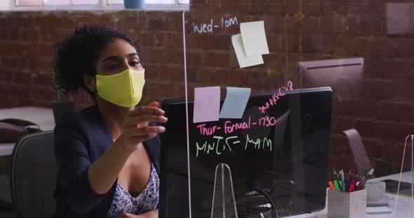 Mixed race businesswoman using memo notes sitting in front of computer wearing face mask