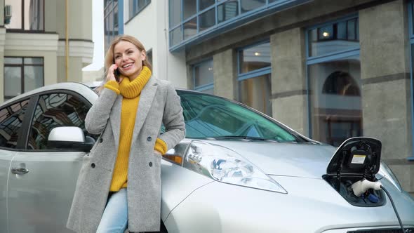 Woman Talking on the Phone While Charging an Electric Car. Environmentally Friendly Transport