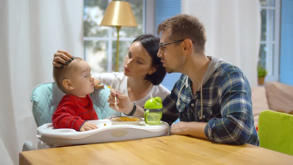 Happy Parents Feeding Baby Son with Healthy Porridge Sitting at Kitchen