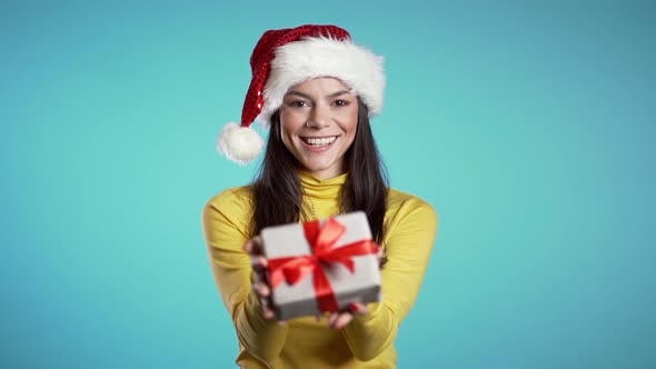 Beautiful cheerful hispanic woman in Santa hat holds out gift box to you on blue background