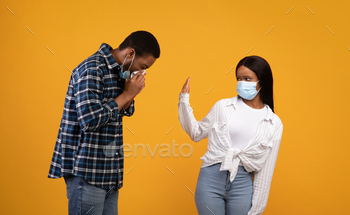 rican lady in protective mask shows stop sign to man, guy sneezes and has first symptoms of disease, isolated on yellow background, studio shot