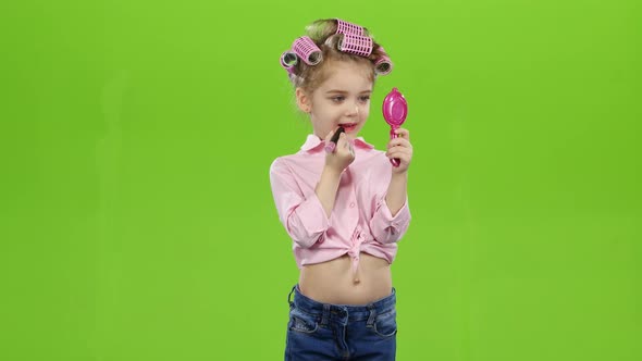 Little Girl in Curlers Holds a Mirror in Her Hands and Paints Lipstick with Lipstick. Green Screen