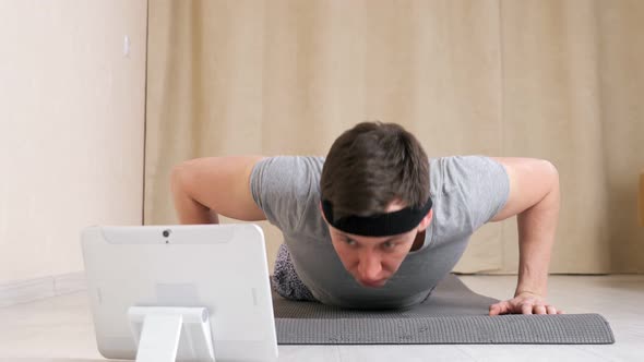 Young Man with a Bandage on His Head Looks Into a Tablet and Clumsily Does Pushups on the Floor