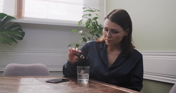 Mature Woman Dripping Drops with Glass of Water, Drinking Sedative Medicine, at Home in Pajamas