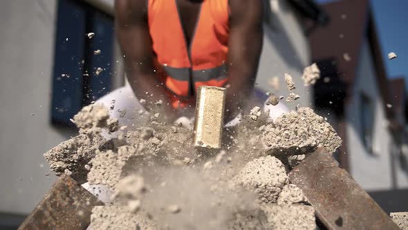 A dark-skinned muscular builder breaks a building stone into small particles with a sledgehammer