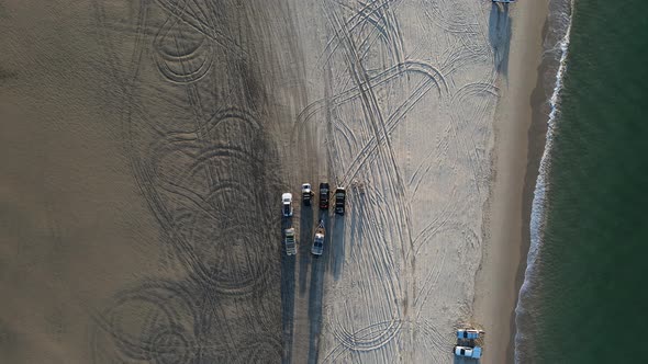 4wd vehicles parked on a beach with the ocean waves lapping the shoreline. Drone view looking down