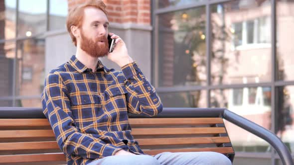 Redhead Beard Young Man Talking on Phone While Sitting Outdoor on Bench