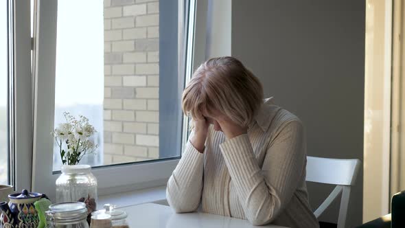 Unhappy Woman Sitting At Table Near Window And Holding Head From Headache