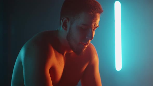 A Tired Athlete is Resting in Breathing Heavily While Sitting on a Barbell in Neon Light After