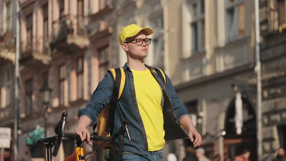 A Young Happy Delivery Man is Going and Holding a Bicycle with Him