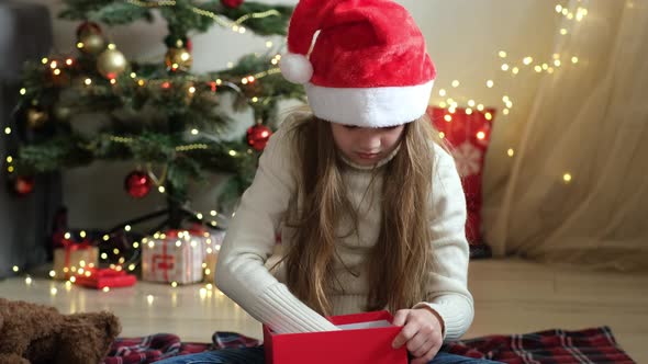 Cute Little Girl in Santa Hat Preparing Christmas Gift on the Background of Decorated Tree at Home