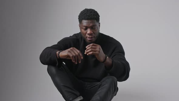 African American Man Covering Face with Hands and Looking Away Sitting in Studio Grey Background