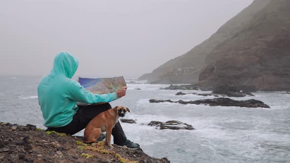 Traveler with Dog Holding Map in Front of Powerful Waves Splashing Into Rocky Coastline