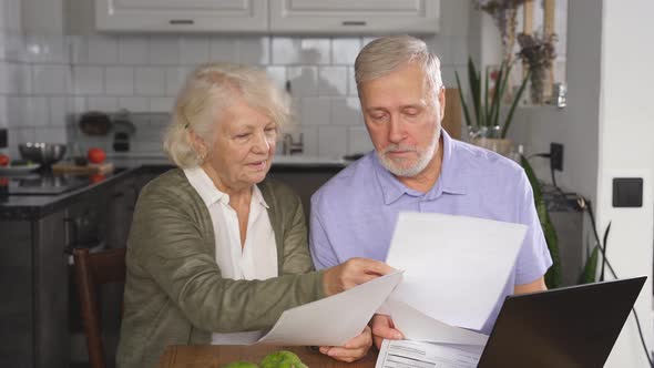 A Sixtyyearold Wife and Husband Sit at a Desk and Laptop Holding Bills in Their Hands Managing the