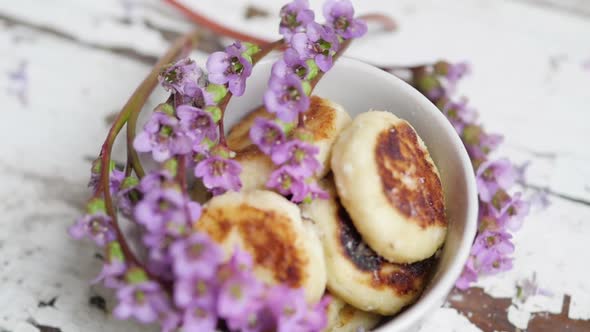 A Bouquet of Lilac Flowers Near a Plate with Cheese Cakes on a Wooden Background