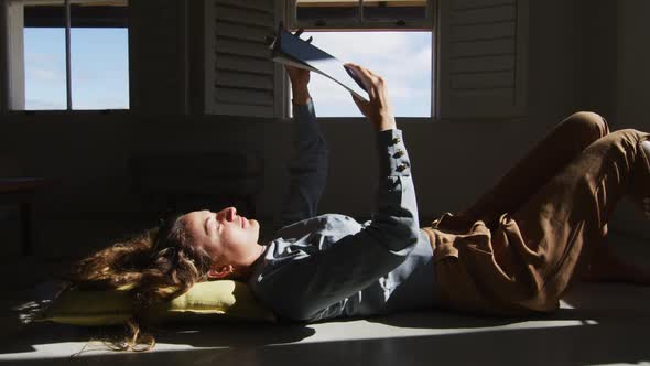 Caucasian woman lying on floor holding book and reading in sunny cottage living room