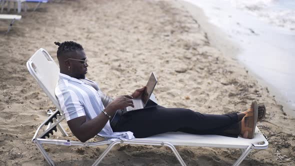 Concentrated Absorbed African American Young Man Lying on Sunbed Surfing Internet on Tablet