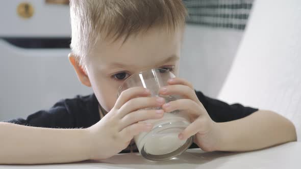 Cute Little Boy Drinking Fresh Milk Into Glass Sitting at the Table in the Kitchen